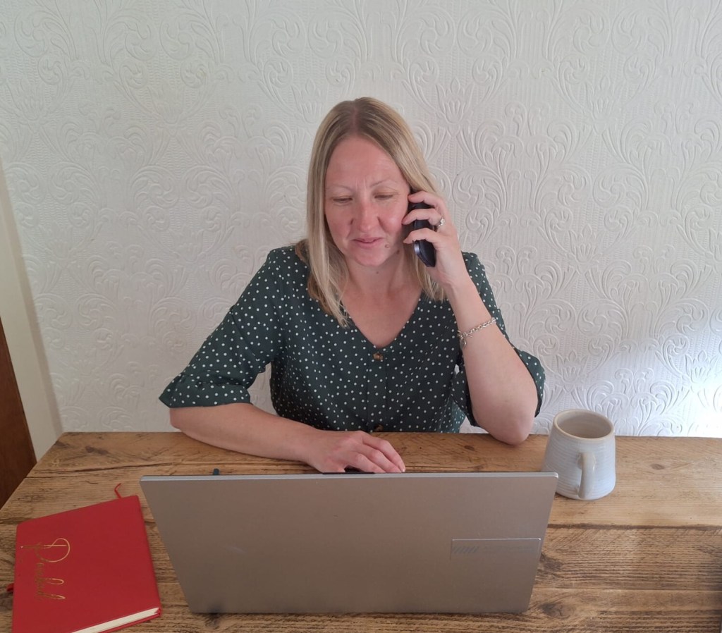 Rosie Mowat, financial coach, speaking with a client over the phone while working on a laptop at a wooden desk with a notebook and coffee cup.