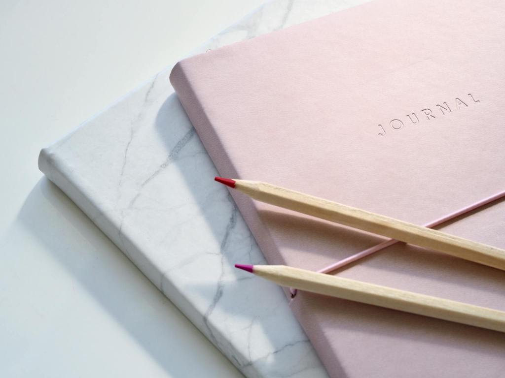 Soft pink and marble journals with gold pencils on a white desk, symbolising reflection and mindful money awareness.